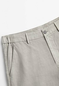 Pantalon gris clair avec passants pour ceinture, poche latérale, fermeture par bouton et une partie élastique à la taille pour un ajustement réglable.