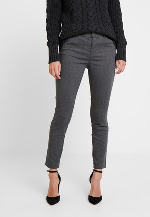 Mujer con pantalones ajustados gris oscuro, suéter de cable negro y tacones altos con correa en el tobillo, de pie contra un fondo blanco.