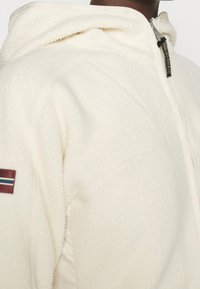 Persona che indossa una giacca in pile color crema con zip anteriore, colletto alto e un piccolo patch con bandiera sul braccio superiore.