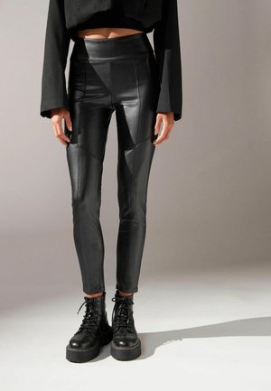 Personne portant des leggings noirs taille haute en cuir synthétique et des bottes noires à lacets épaisses, debout devant un fond clair uni.