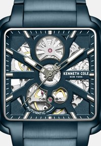 Quadratische Automatikuhr mit einem blauen Edelstahlgehäuse und -armband. Transparenter Ziffernblatt zeigt filigrane Zahnräder und silberne Akzente.