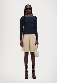 Marineblå langermet topp med firkantet utringning, beige skreddersydde shorts, burgunder ankelstøvler og en kremfarget håndveske; modell med langt fletthår.