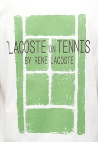 Witte T-shirt met een graphic van groene rechthoeken die lijken op een tennisbaan; tekst luidt "LACOSTE on TENNIS BY RENE LACOSTE" in het zwart.