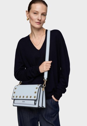 Mujer que lleva pendientes de aro dorados y suéter negro de cuello en V, sosteniendo un bolso de cuero azul claro con ojetes dorados y correa ancha.