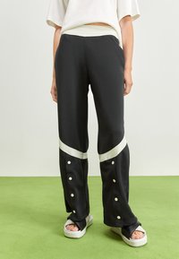 Person klädd i svarta träningsbyxor med vita diagonala ränder och knappsydd nederdel, tillsammans med svarta och vita sandaler.