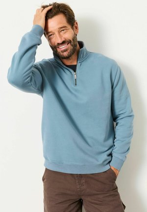 Smiling man with beard wearing blue quarter-zip sweatshirt and brown pants, running hand through hair against white background.