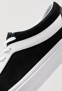 Černé semišové tenisky s bílým koženým bočním pruhem, bílými tkaničkami a silnou bílou gumovou podrážkou na neutrálním pozadí.