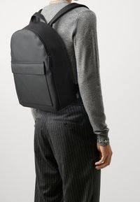 Svart ryggsäck med en slät, matt finish; har en framficka, rundad topp och justerbara remmar. Bärs över en grå tröja med randiga byxor.