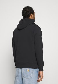 Felpa nera in misto cotone, con un ampio cappuccio e polsini a coste. Chi indossa è di spalle, con dei jeans blu chiaro visibili.