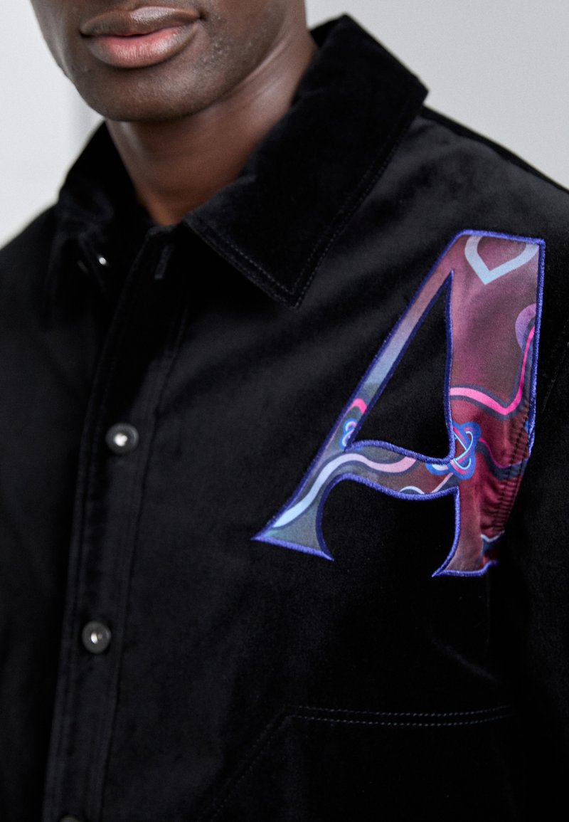 Schwarzes, samtiges Hemd mit einem großen, bunten bestickten Buchstaben "A" und wirbelnden Mustern, das das elegante Design und die Knopfdetails ergänzt.