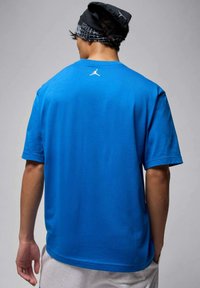 Blaues Baumwoll-T-Shirt mit kurzen Ärmeln, versehen mit einem weißen Jumpman-Logo am Hals und einer lockeren Passform. Getragen mit einem schwarzen, gemusterten Kopftuch.