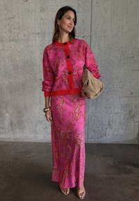 Γυναίκα με ρόζ ζακέτα με παίζεϊλι σχέδιο και μακριά φούστα που ταιριάζει, κρατώντας μπεζ τσαντάκι, μπροστά σε τσιμεντένιο τοίχο.