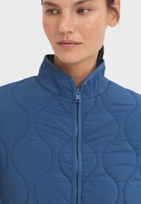 Veste matelassée bleue avec un col montant et une fermeture éclair, présentant un motif en vagues et une texture lisse.