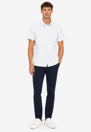 Mann trägt ein weißes kurzärmliges Hemd mit kleinem dunklem Muster, marineblaue Hose und weiße Sneakers, steht mit einer Hand in der Tasche.