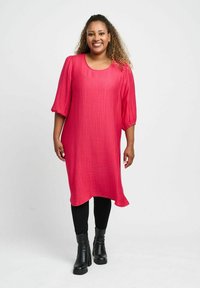 Rosa, strukturiertes Tunika-Kleid mit lässiger Passform und dreiviertel Ärmeln; kombiniert mit schwarzen Leggings und Stiefeletten.