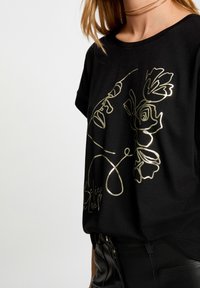 Camiseta negra de manga corta con un diseño floral en oro metálico y texto en relieve en el dobladillo. Tela suave con un corte relajado.