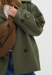 Giacca oversized verde oliva con grandi bottoni neri e colletto ampio, abbinata a una borsa a tracolla in tessuto morbido marrone.
