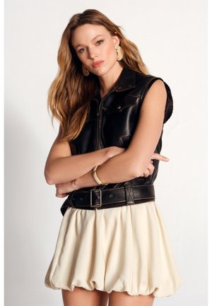 Donna con capelli lunghi e ondulati che indossa grandi orecchini d’oro, un gilet nero in pelle senza maniche con cintura e una gonna corta color crema, braccia incrociate.