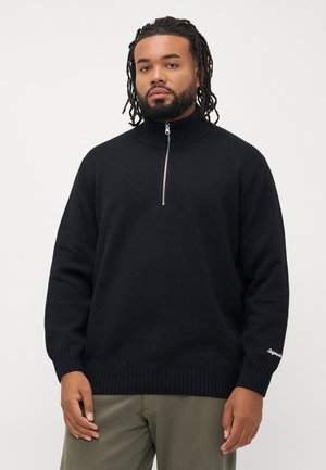 Pull en tricot noir avec un col montant, fermeture éclair moitié, poignets et ourlet côtelés, portant un texte brodé sur la manche.