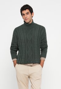 Homem vestindo um suéter de gola alta em ponto cabeludo verde escuro sobre uma camisa xadrez e calças beges, com as mãos nos bolsos, contra um fundo simples.