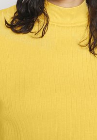 Žlutý žebrovaný pletený top s vysokým límcem. Látka je texturovaná, vykazuje vertikální pruhy a těsný střih.