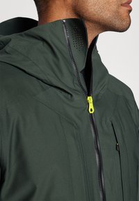 Homme portant une veste à capuche vert foncé avec une tirette jaune néon, veste zippée près du menton, montrant le profil latéral du cou et de la mâchoire.