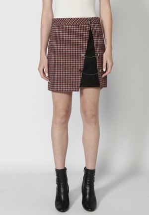 Jupe mini en tartan rouge, blanc et noir avec une taille haute, ouverture avant sécurisée par des boutons et accents en chaîne sur le côté.