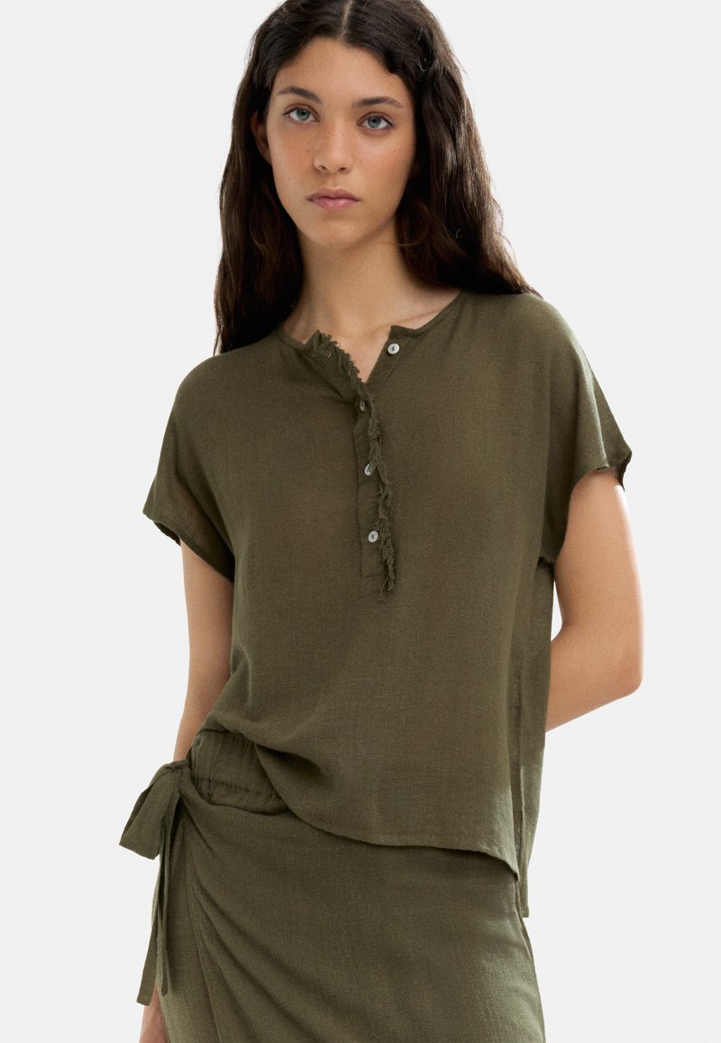 Oliwkowa, lekka bluzka z okrągłym dekoltem, strzępieniami i krótkimi rękawami, z listwą guzikową i luźnym krojem.