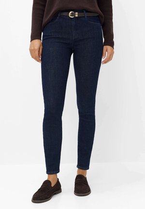 Person iført mørkeblå skinny jeans, sort bælte med guldspænde, brun sweater og mørkebrun ruskindsløber, stående mod hvid baggrund.