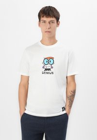 Ung mand iført en hvid T-shirt med en tegneseriefigur med store blå briller og tekstet "GENIUS", mørke bukser og en tatovering på sin underarm.