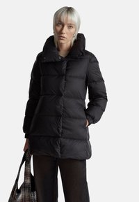 Černá péřová bunda s vysokým límcem, prošívaným designem a cvočky. Modelka drží vzorovanou tašku a má na sobě tmavé kalhoty.