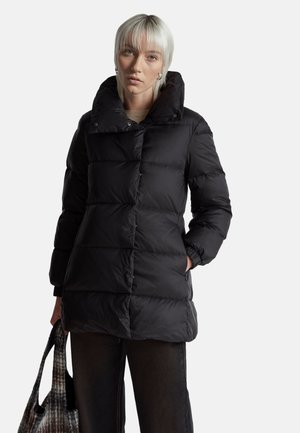 Schwarze Pufferjacke mit hohem Kragen, gesteppter Gestaltung und Druckknopfverschlüssen. Das Modell hält eine gemusterte Tragetasche und trägt dunkle Hosen.