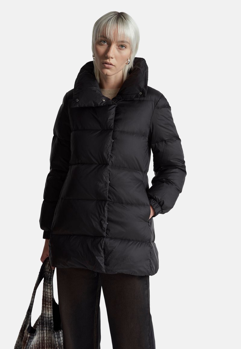 Černá péřová bunda s vysokým límcem, prošívaným designem a cvočky. Modelka drží vzorovanou tašku a má na sobě tmavé kalhoty.