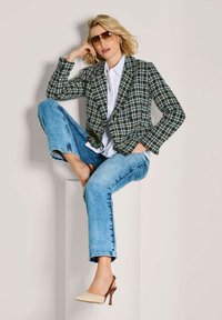 Frau mit kurzem blonden Haar, die Sonnenbrillen trägt, einen karierten Blazer, ein weißes Hemd, blaue Jeans und beigen Absätzen, sitzt auf einem weißen Block.