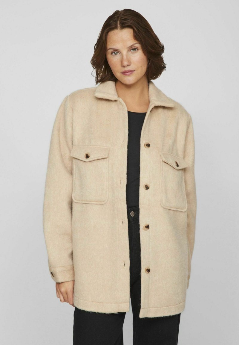 Beige villasekoite takki, jossa on kauluksellinen kaulus, napitus ja kaksi etutaskua. Pehmeä rakenne ja hieman ylisuuri istuvuus.