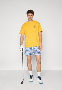 Rumena bombažna majica s potiskom, v paru z modrimi kratkimi hlačami z geometrijskimi vzorci. Bele teniske in golf palica v roki.
