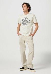 Vit t-shirt med kort ärm och tryckt svart text, parat med ljusbeiga byxor och svarta sneakers med krämfärgade detaljer.