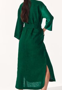 Długa zielona lniana sukienka z paskiem, rękawami o długości trzech ćwiartek i bocznymi rozcięciami. Materiał ma fakturowane, matowe wykończenie.