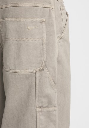 Béžové nohavice typu cargo s textúrovanou tkaninou, s bočnou vreckom a malým vyšívaným logom Nike na zadnom vrecku.