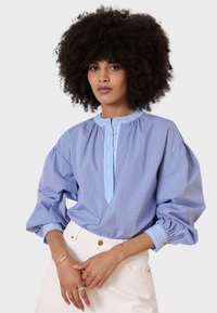 Camicia a quadri blu e bianca con maniche a sbuffo, colletto a stand e placket con bottoni. Accenti blu chiaro sul colletto e sui polsini.