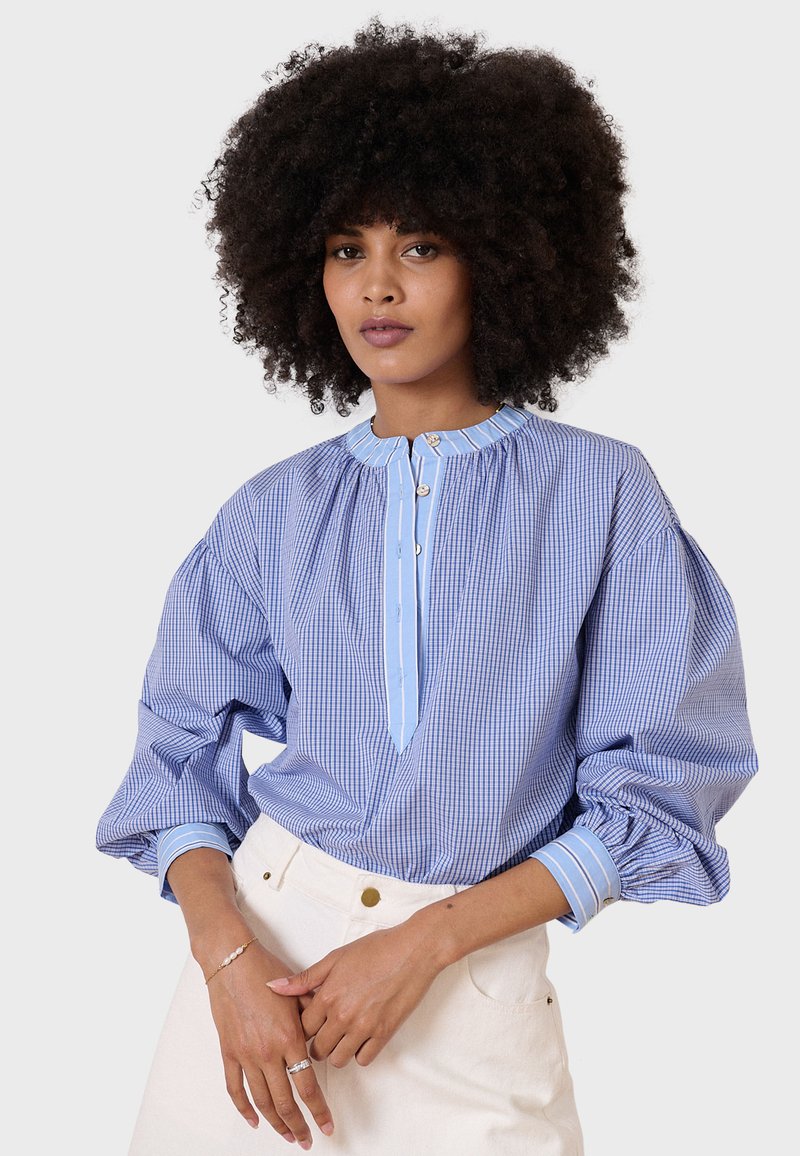 Camicia a quadri blu e bianca con maniche a sbuffo, colletto a stand e placket con bottoni. Accenti blu chiaro sul colletto e sui polsini.