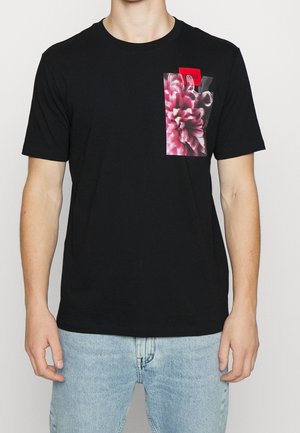 T-shirt nera in cotone con maniche corte, con una stampa di una fiore rosa sulla tasca frontale e un'etichetta rossa. Design semplice e casual.