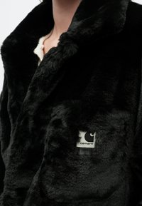 Chaqueta de piel sintética negra con cuello de capa, que cuenta con un bolsillo frontal y una etiqueta con el logo de Carhartt. Textura suave y apariencia esponjosa.
