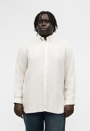 Sötét bőrű személy rövid dreadlock frizurával, világos bézs színű, hosszú ujjú, gombos ingben és sötétkék nadrágban, egyszínű háttér előtt állva.