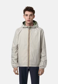 Giacca impermeabile grigio chiaro con cappuccio, chiusura con zip e polsini elasticizzati. Presenta una fodera verde e un dettaglio della zip multicolore.