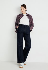 Pantaloni in denim scuro a gamba larga con pieghe frontali, abbinati a una maglietta bianca e una camicia a righe bordeaux con bottoni, e sneakers chiare.
