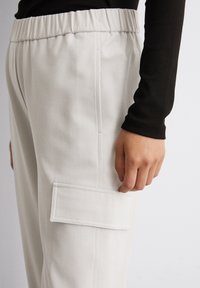 Helle graue Jogginghosen mit strukturierter Oberfläche, elastischem Bund, Seitentasche und aufgesetzter Tasche am Oberschenkel, kombiniert mit einem schwarzen Langarmshirt.