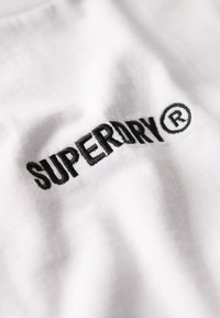 Hvid bomulds t-shirt med fremtrædende sort broderet tekst "SUPERDRY®" på brystet, med en glat tekstur og subtil glans.