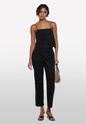 Top nero con ricami floreali e spalline sottili, abbinato a pantaloni neri con motivi a taglio e una borsa intrecciata. Tacchi aperti neri.