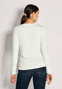 Frau mit dunklem Haar im Pferdeschwanz, die ein weißes Langarmshirt und dunkelblaue Jeans trägt, steht an einer hellen Wand.